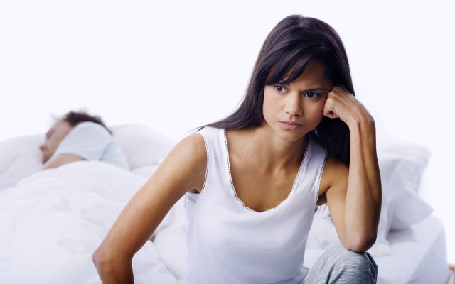 Frustrated woman sitting at edge of bed while her boyfriend sleeps